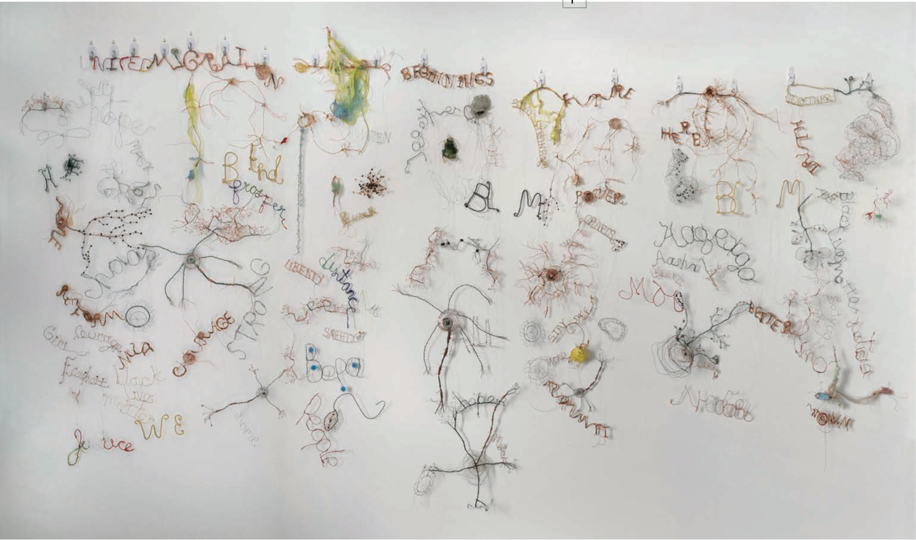 Image Description: A white wall with approximately 50 hand-twisted wire pieces hanging next to one another in a scattered manner. The pieces are made of various metals like copper, silver, and gold, and notable pieces read words like "BLM," "Courage," and "Strong." Some wire pieces are bent into more abstract shapes and forms.